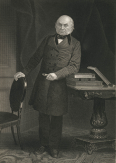 A white man in a long coat stands next to a desk.